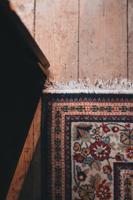 A detailed view of a traditional decorative rug with intricate floral patterns in red, blue, beige, and green, situated on a wooden floor with visible natural grain and a honey-colored finish. The rug