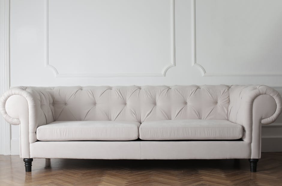 A person dressed in a dark jacket uses a black handheld vacuum cleaner to deep clean a decorative yellow and white patterned cushion placed on a dark fabric sofa in a living room. The scene is illumin