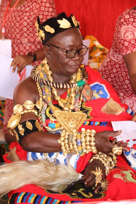 A woman dressed in traditional Maasai attire, adorned with elaborate gold jewelry and beaded accessories, is seated indoors on a red and patterned fabric surface. The background features red patterned